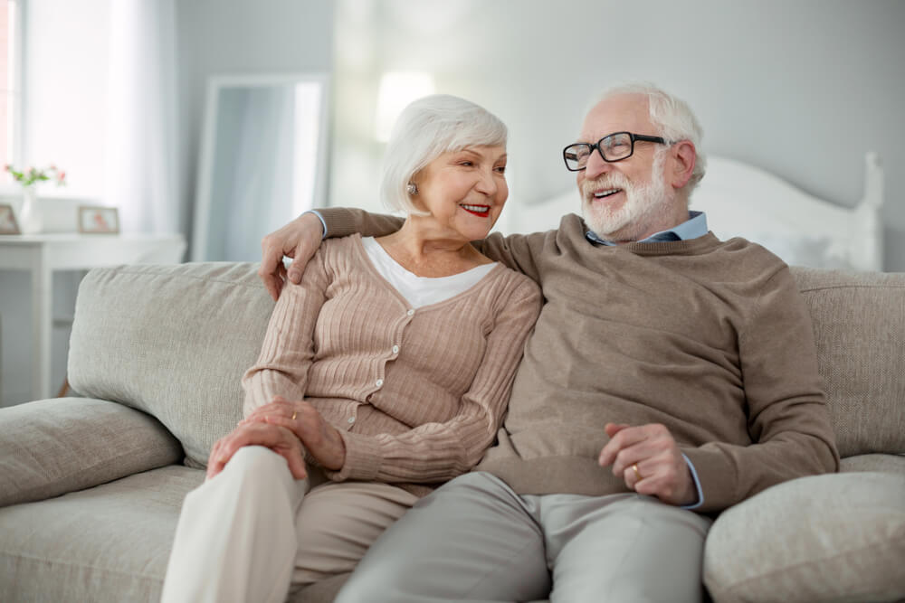 A joyful elderly pair basks in mutual comfort, nestled on a sofa within their senior living community apartment. Enjoying retirement life while sharing heartfelt smiles and companionship.