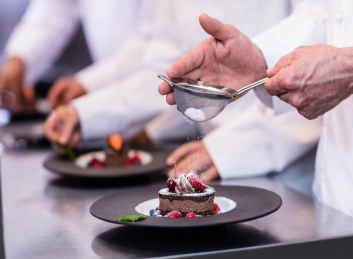 Renowned chef expertly adding a finishing touch of powdered sugar to a dessert at our retirement community's dining hall.