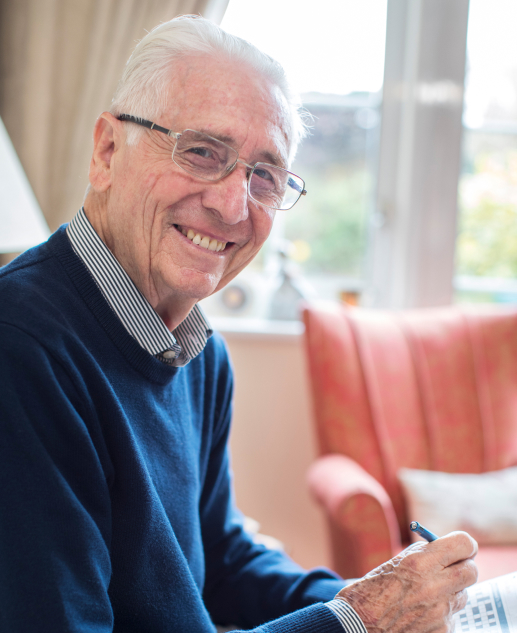 Delighted senior gentleman, sporting glasses and brandishing a pen, posing for the camera by the window of his comfortable living space at Mira Vie at Brookfield Retirement Community.