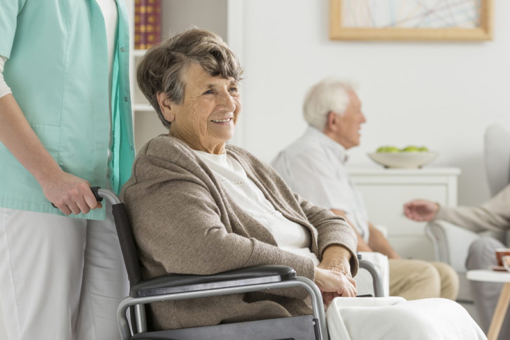 Smiling senior woman in a wheelchair, assisted by our dedicated healthcare worker, with another community member enjoying the scenic backdrop of our retirement home.