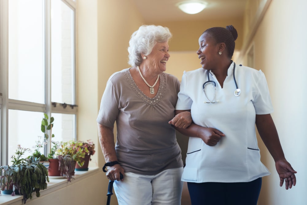 Senior woman using a cane strolling with a cheerful nurse in a retirement home corridor.