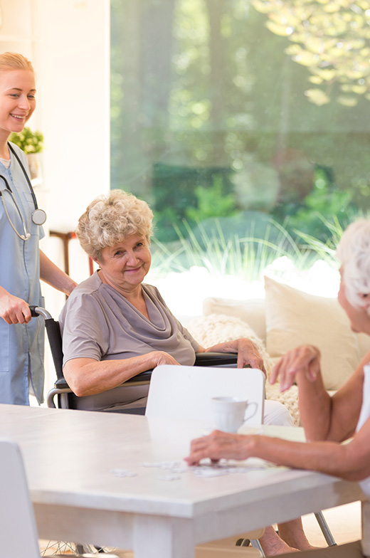 An elderly lady smiles while in the comfort of our memory care facility, enjoying quality social time with another senior resident. A professional nurse stands watch to ensure safety and support. Our bright, cozy environment ensures a comfortable retirement lifestyle for all seniors at our home.