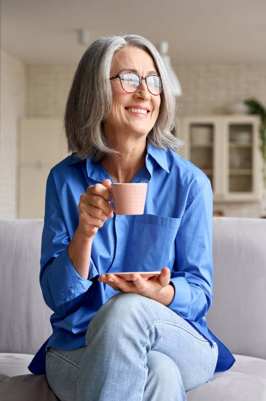 A cheerful elderly woman, clad in a blue shirt, enjoys her coffee on a couch within our senior living community.