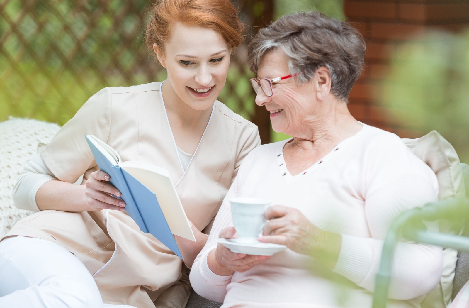 A beautiful, red-headed nurse reads to a memory care resident on the patio.