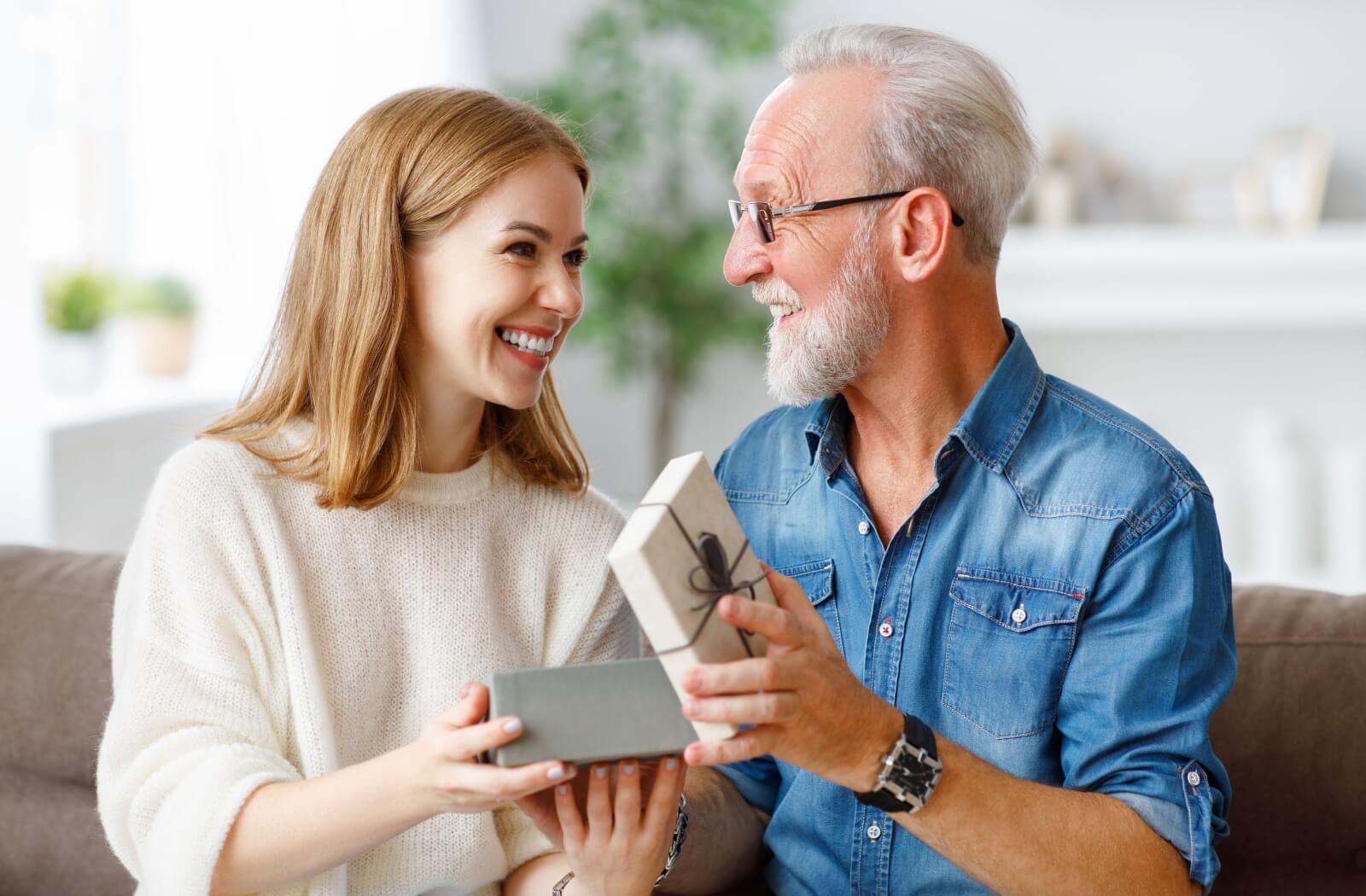 An older adult smiling while exchanging a gift with a younger adult in a cozy living room setting.