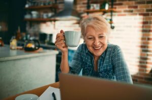 An older adult in a kitchen raising a mug and smiling while looking at a laptop.