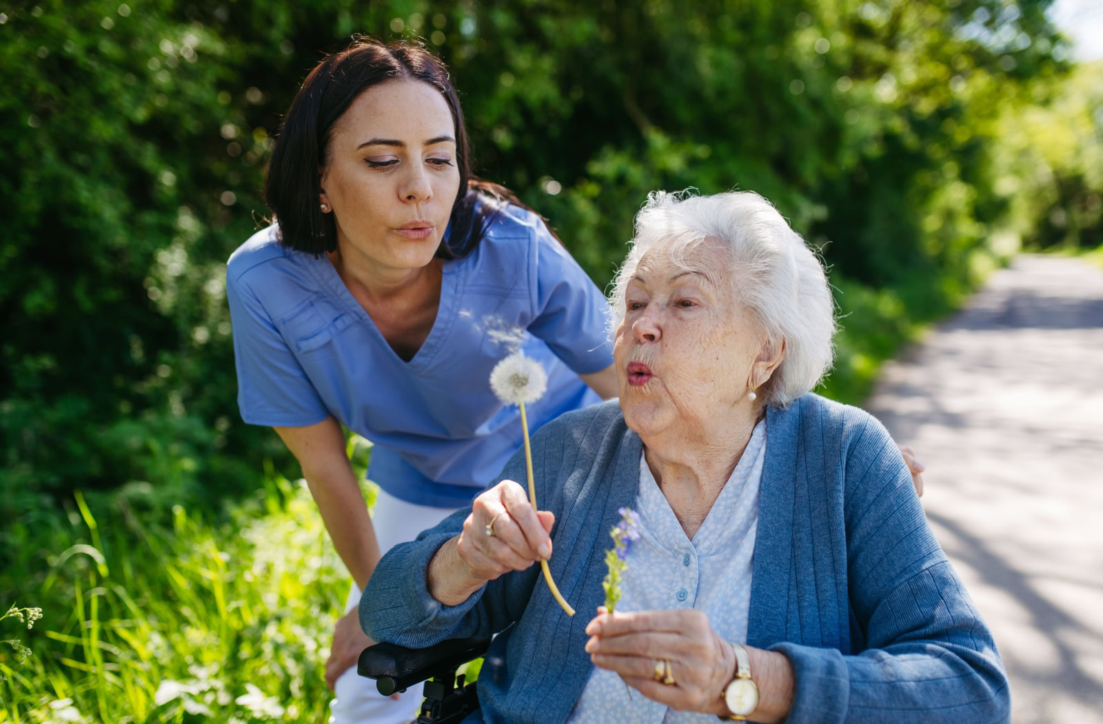 Caretaker and assisted living resident in a wheelchair are outside together, both blowing on a dandelion.