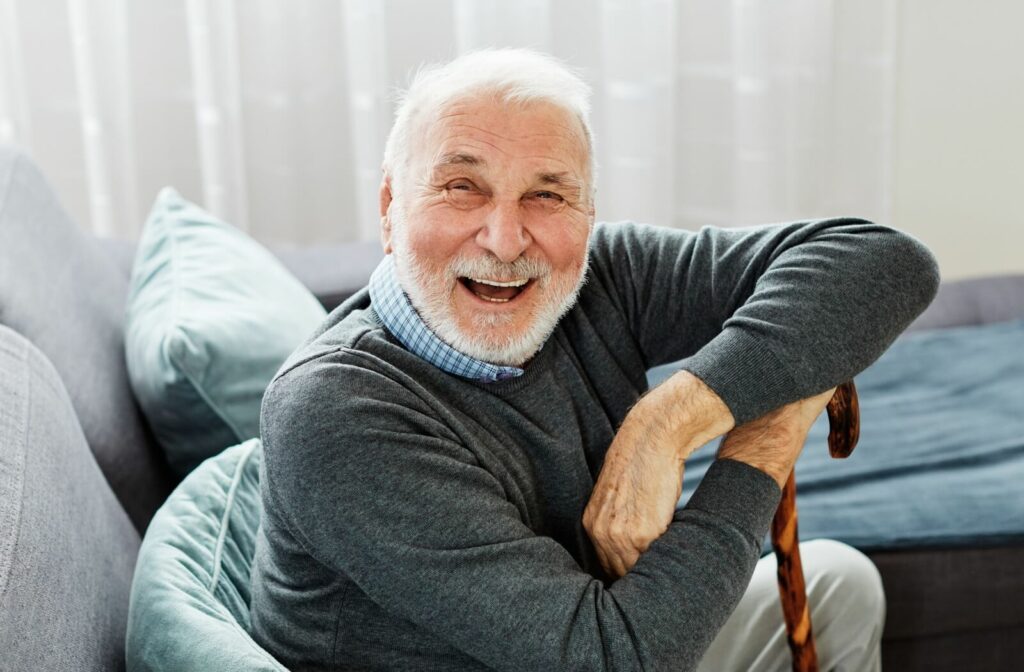 A smiling older adult sitting on a sofa, holding a cane, in a bright and cozy living room