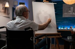 An older adult in a wheelchair sketching a still life on an easel in a warmly lit room.