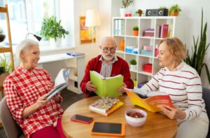 Three seniors sit at a circular table in a bright room, holding different books open and talking about what they’re reading
