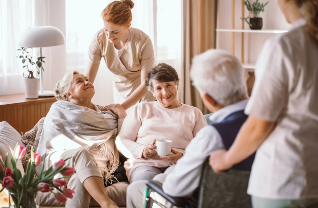 A trio of seniors socializing in a warm common area with plants are comforted by nurses in a memory care community