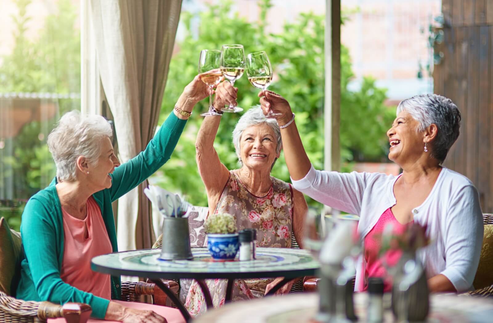Three seniors clink together glasses of white wine while they sit at an outdoor patio table, enjoying each other’s company