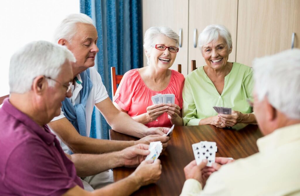 A group of smiling seniors plays a game of cards at a table in an assisted living community.