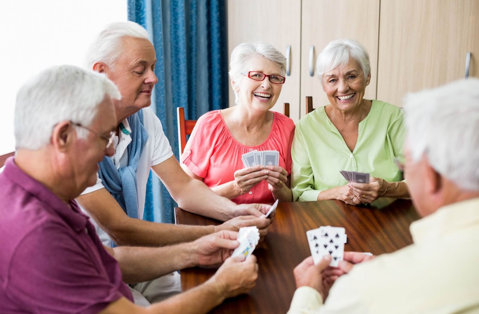 A group of smiling seniors plays a game of cards at a table in an assisted living community.
