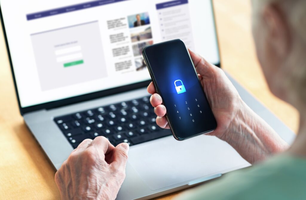 An older adult browsing social media on a laptop holds up their locked phone.