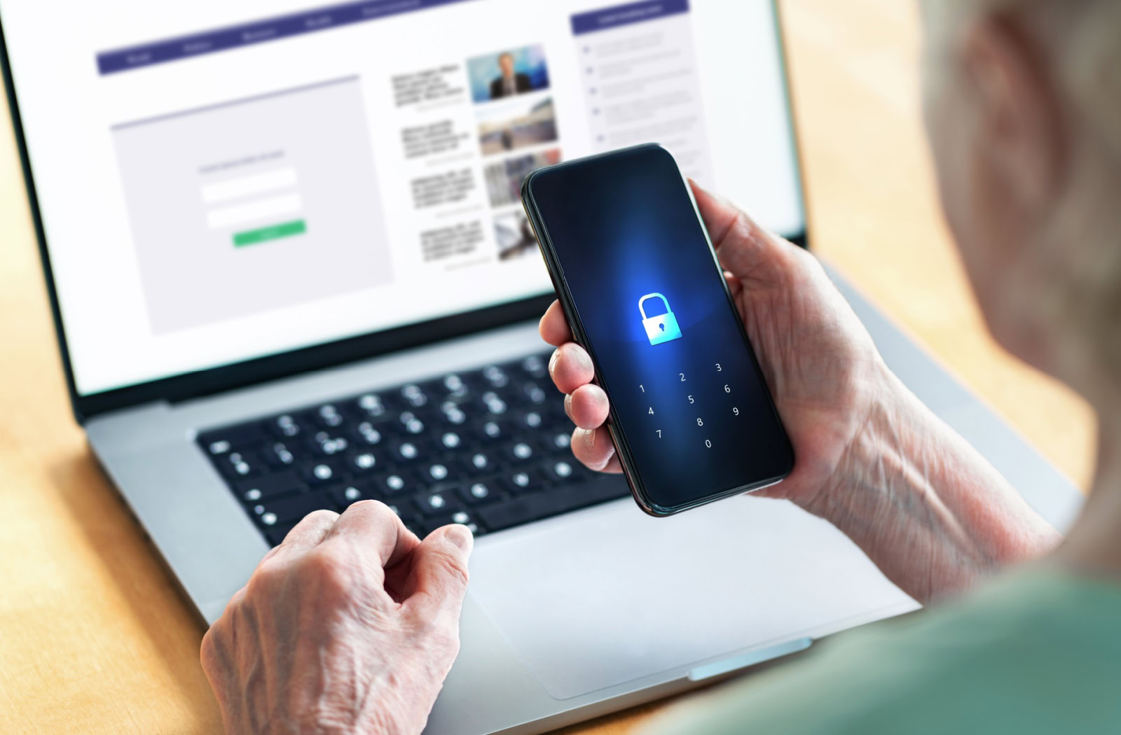 An older adult browsing social media on a laptop holds up their locked phone.