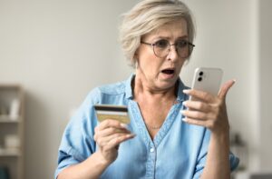 An older adult looks at their phone in shock as they hold their credit card in their other hand.
