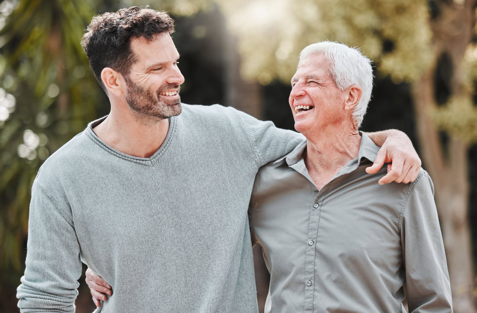 A smiling senior and their adult child chat and enjoy a nice walk in the senior community with their arms around one another's shoulder.