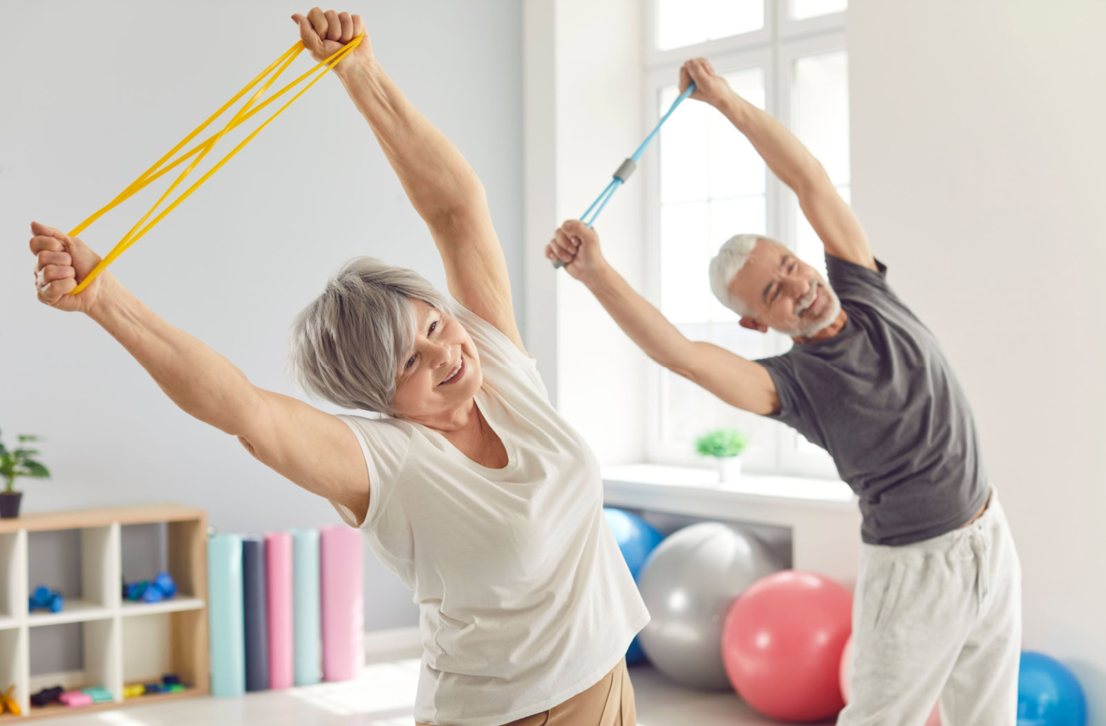 A senior couple each hold exercise bands over their heads as they stretch to the side during an exercise class