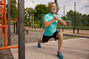 A senior performs a lunge while holding their hands together at an outdoor park with exercise equipment for adults