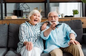 a senior couple sit on a couch laughing at a move on their tv