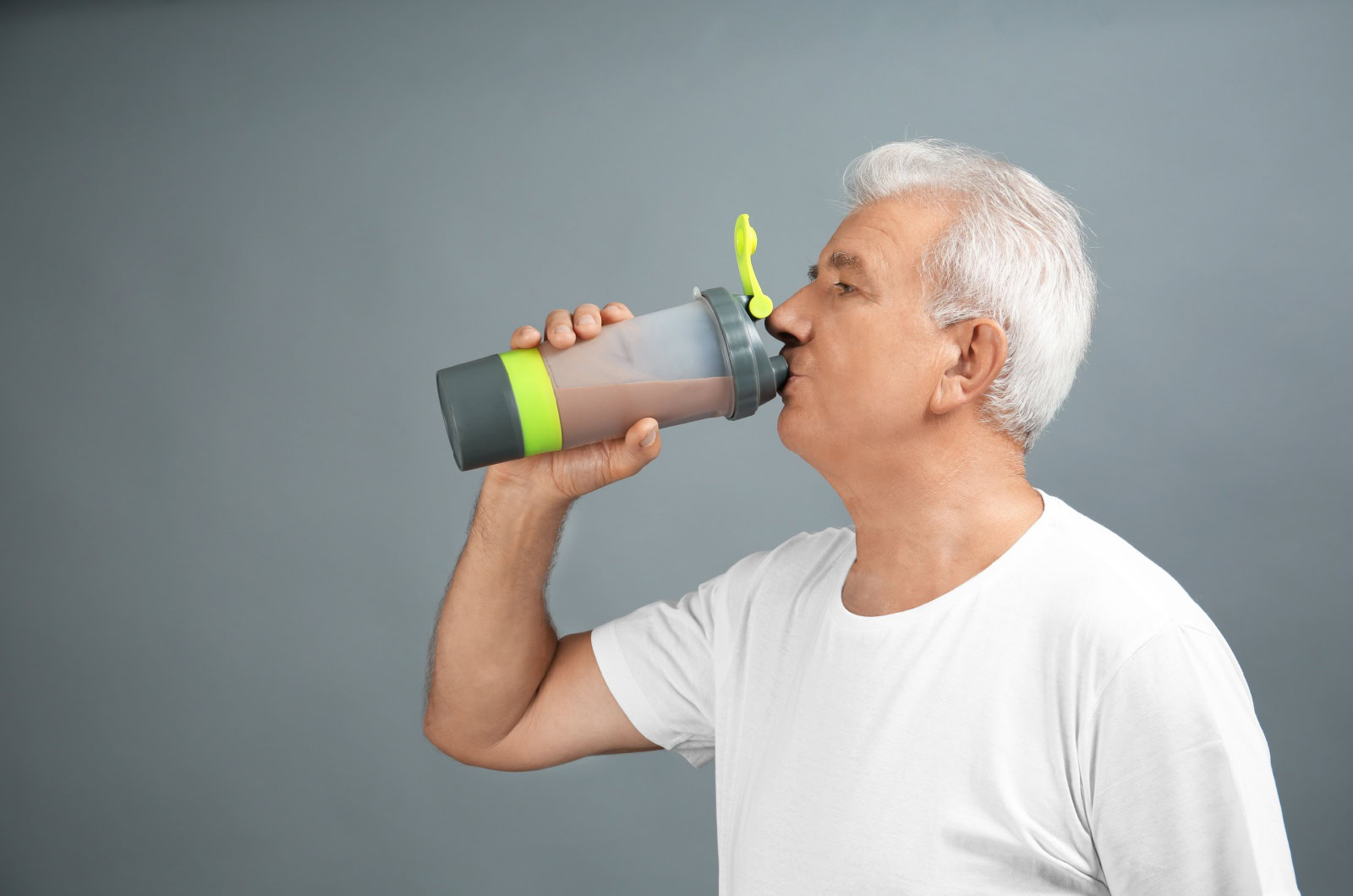 n older adult in a white t-shirt drinking a protein shake from a bottle in his right hand.