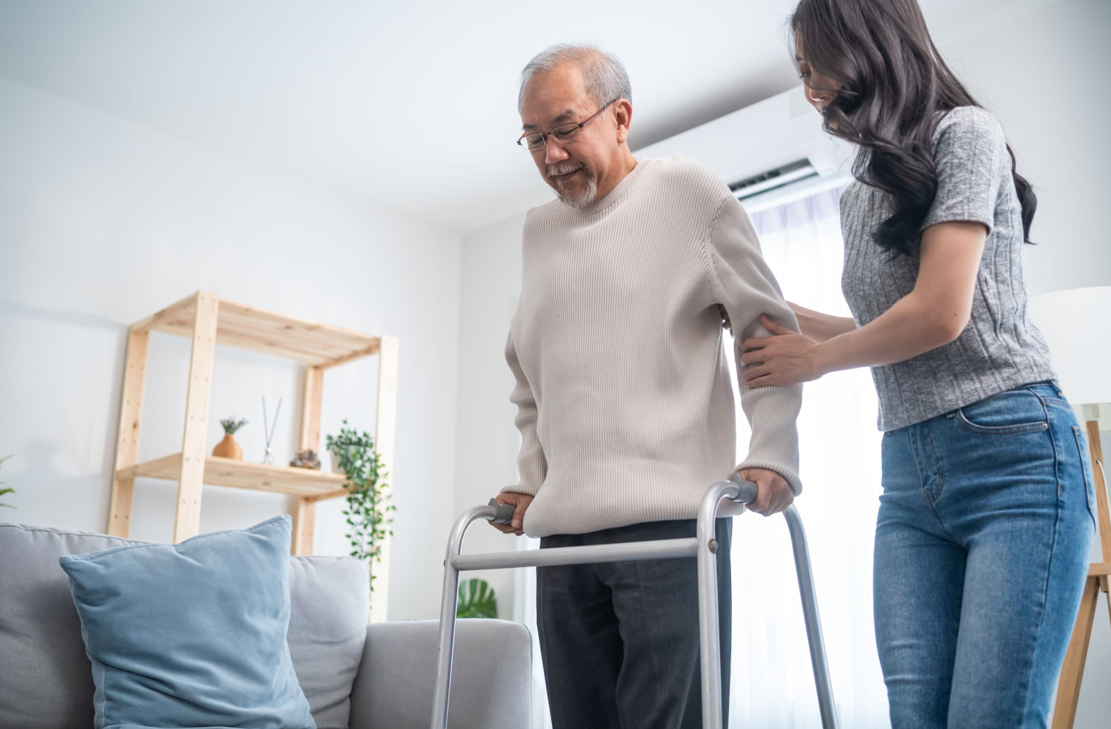 An older adult using a walker with assistance from a caregiver in a bright and cozy living room.