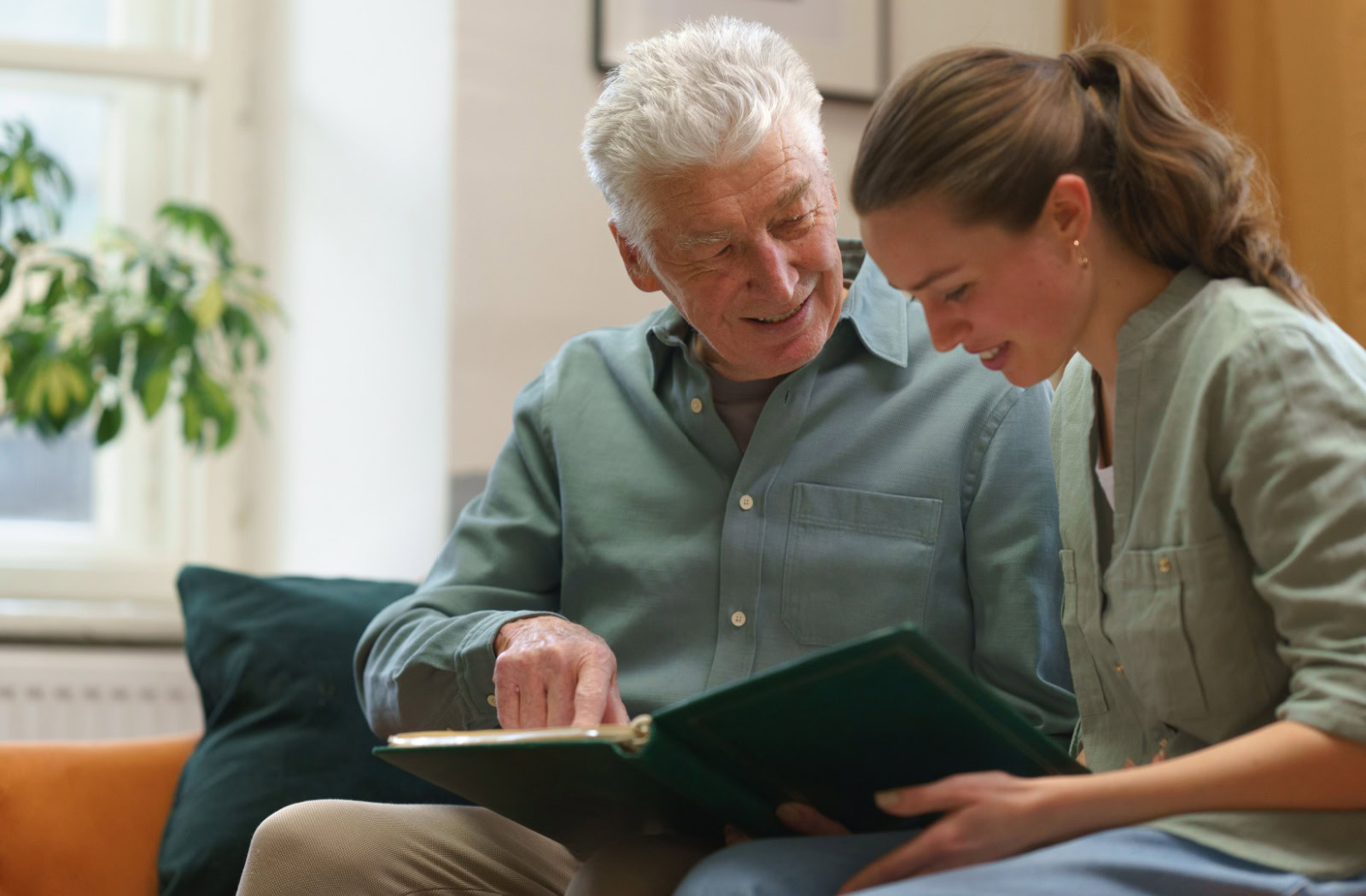 n elderly man and daughter sitting together on a couch, smiling as they look through a photo album.