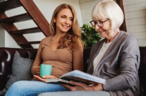 A woman holding a mug, smiling warmly at her mother, who is reading a book as they sit together on a cozy couch.