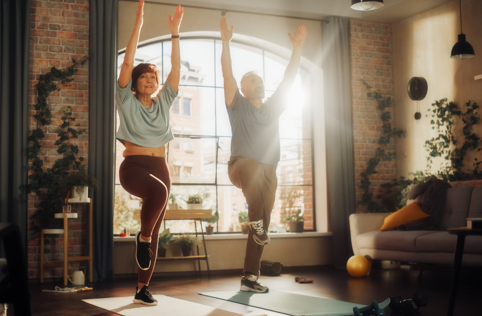 A senior couple each balance on one leg while raising their arms in the living room of their trendy apartment