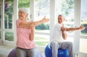 A senior couple each balance on exercise balls while stretching an arm over their chest as part of morning exercise