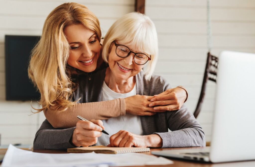 An adult child hugging their older parent from behind while helping them sign legal documents.