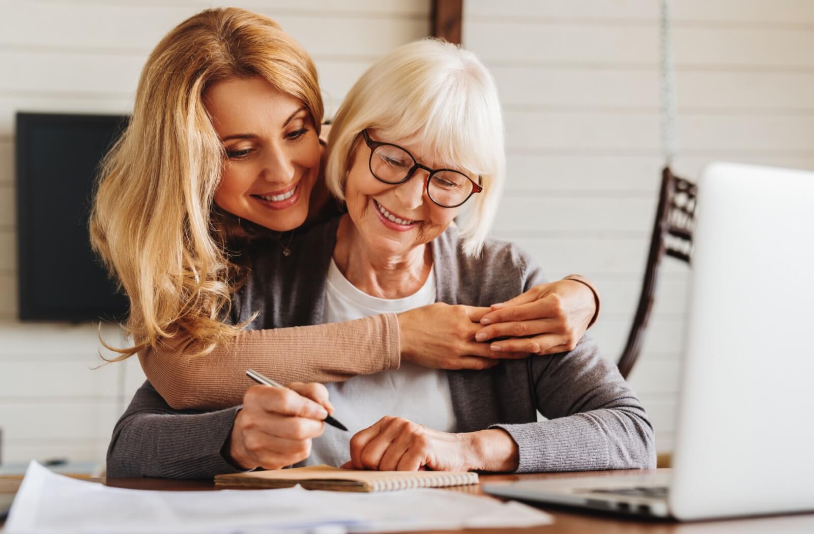 An adult child hugging their older parent from behind while helping them sign legal documents.