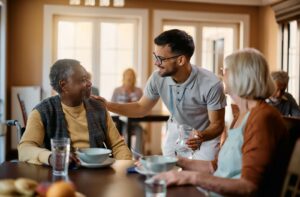 A caregiver smiling while checking in on two residents in assisted living during breakfast.