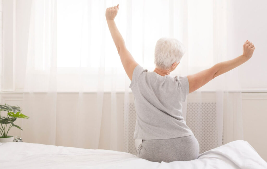 A senior sits facing the window at the edge of their bed in the morning. They raise their arms in a large stretch.