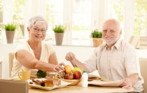Two seniors sit in a bright kitchen, eating their nutritious breakfast of toast, fruit, and bacon.