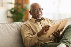 An older adult sitting cross-legged on their couch in senior living while reading a book about Alzheimer's disease.