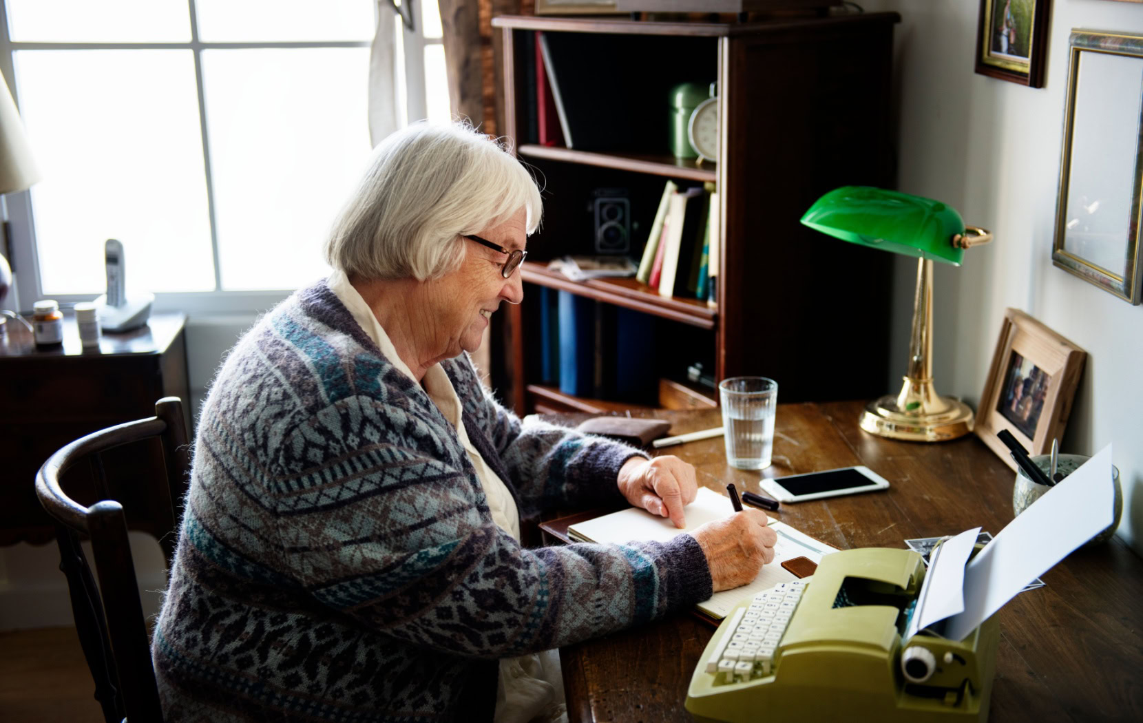 A senior sits at their desk in their home office and writes in a gratitude journal while smiling softly to themselves.