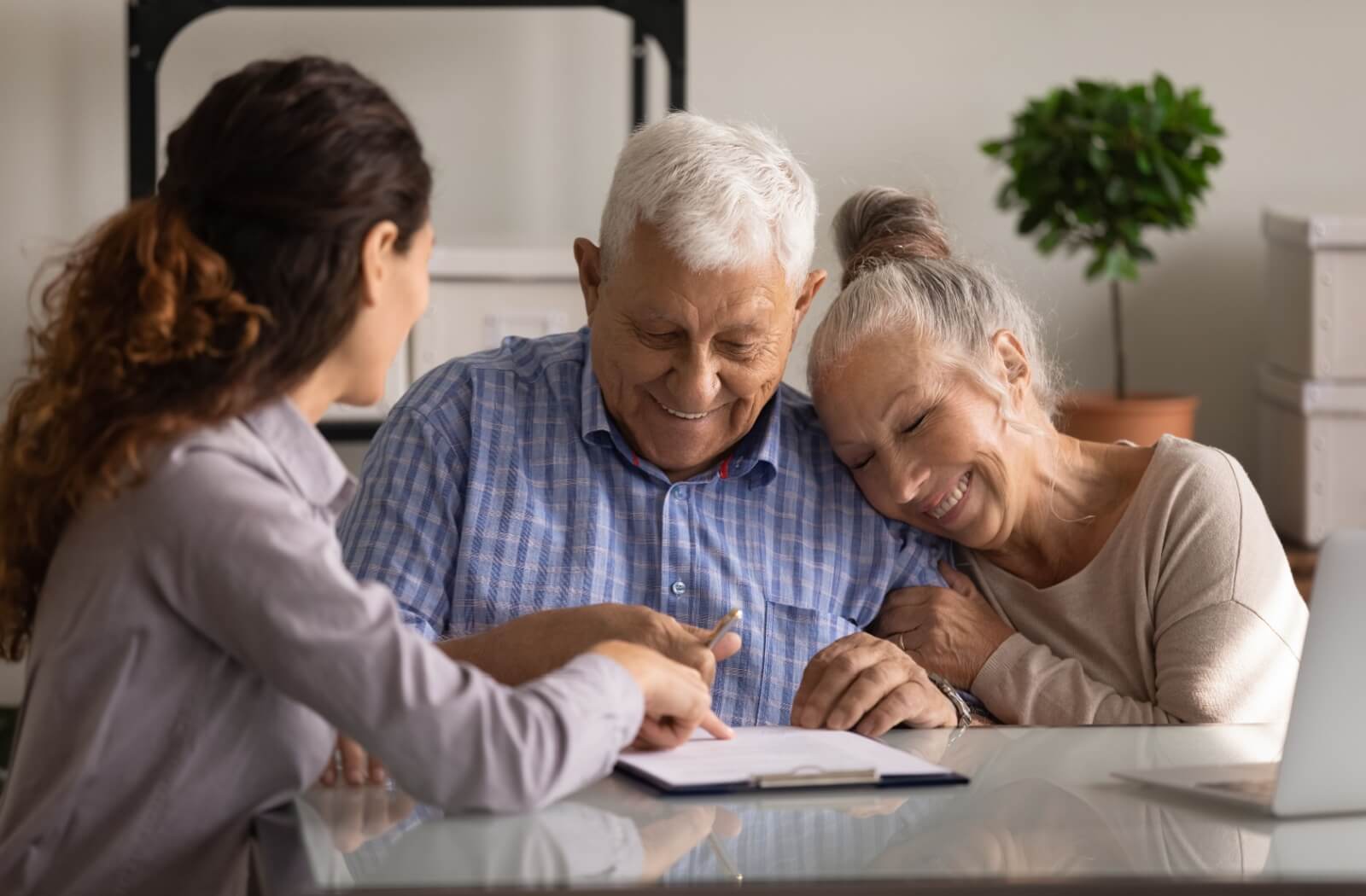 An older adult with dementia signing a legal document for guardianship while their partner hugs them and a caregiver helps.