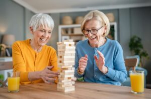 2 older friends indoors in senior living, drinking juice and playing a fun game of Jenga together.