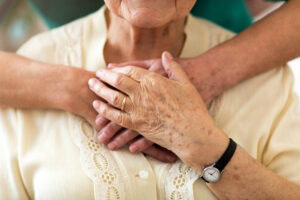A senior clasps a loved one's hands while being embraced from behind.