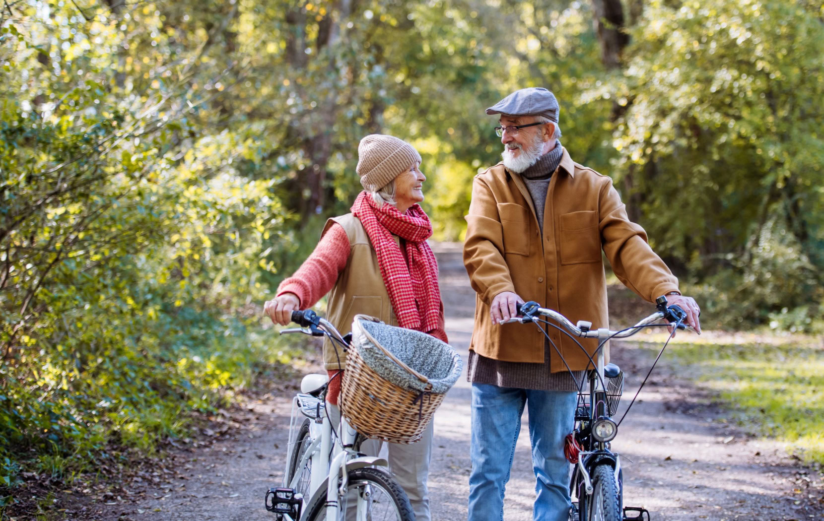 A senior couple wearing warm jackets chat as they walk their bikes down a park path on a crisp autumn morning.
