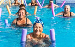 A group of older adults use pool noodles during a low-impact water aerobics class at their senior living community.