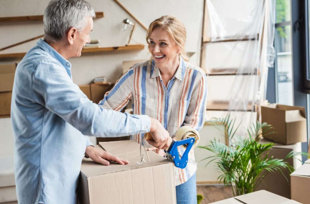 An adult helping their older parent tape up a moving box while packing their home to downsize for assisted living