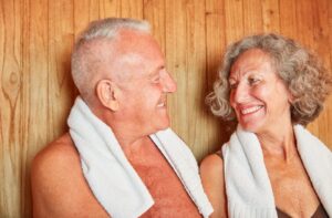 A senior couple with towels around their necks smiling at each other inside a wooden sauna.