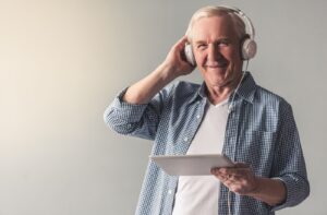 A grinning older adult holding a tablet and wearing headphones while listening to a history podcast.
