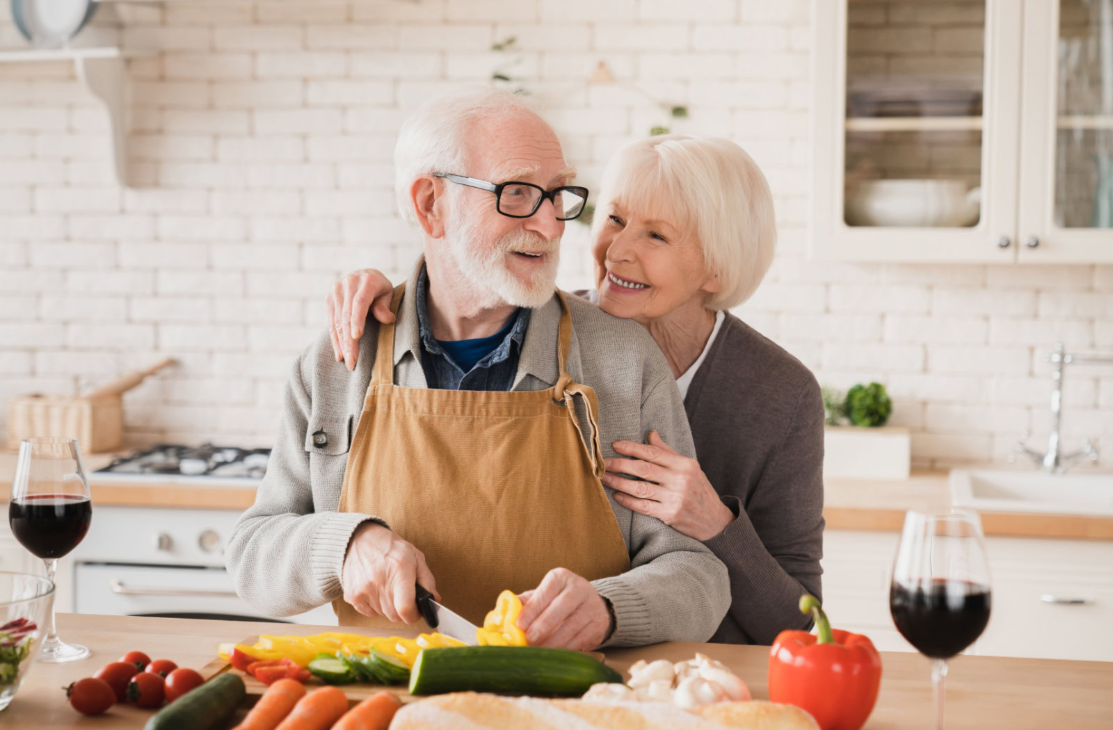 An older married couple smiling and hugging in the kitchen while working together to chop vegetables.