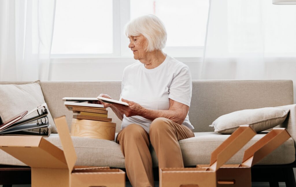 A senior sits on their couch with open moving boxes at their feet. They are looking through old photos and items and deciding what to keep and what to declutter.