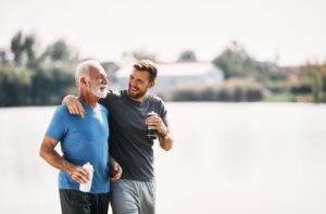 An adult child with their arm around their older parent while taking a break during a jog beside a lake together.
