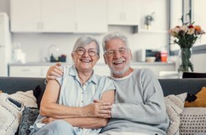 An older couple smiling and sitting on their couch in independent living while hugging.